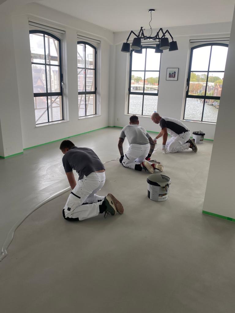Drie stukadoors knielend aan het werk op een gladde vloer in een loft met grote boograamen